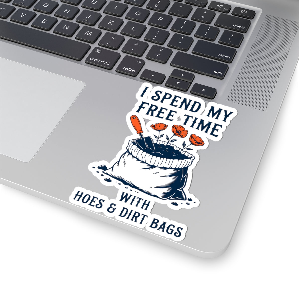 A kiss-cut vinyl sticker applied to the corner of a silver laptop. The sticker features a humorous graphic with the text 'I SPEND MY FREE TIME WITH HOES & DIRT BAGS' surrounding an illustration of a black-and-white bag of dirt with orange gardening tools and flowers.