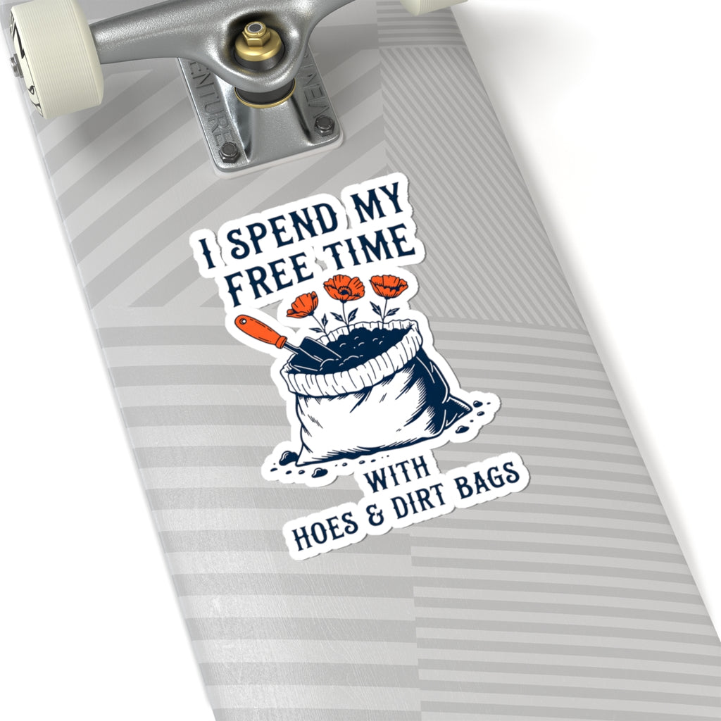 A close-up of a skateboard deck featuring a kiss-cut sticker with the text 'I SPEND MY FREE TIME WITH HOES & DIRT BAGS' and an illustration of a bag of soil with flowers and a trowel. The skateboard has a gray and white striped pattern.