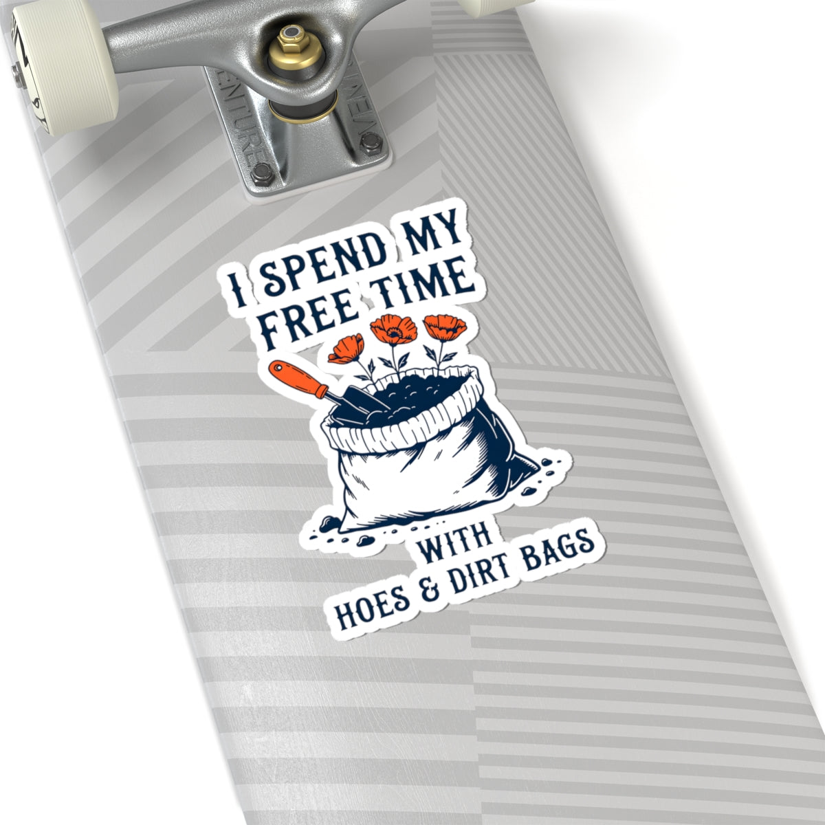 A close-up of a skateboard deck featuring a kiss-cut sticker with the text 'I SPEND MY FREE TIME WITH HOES & DIRT BAGS' and an illustration of a bag of soil with flowers and a trowel. The skateboard has a gray and white striped pattern.