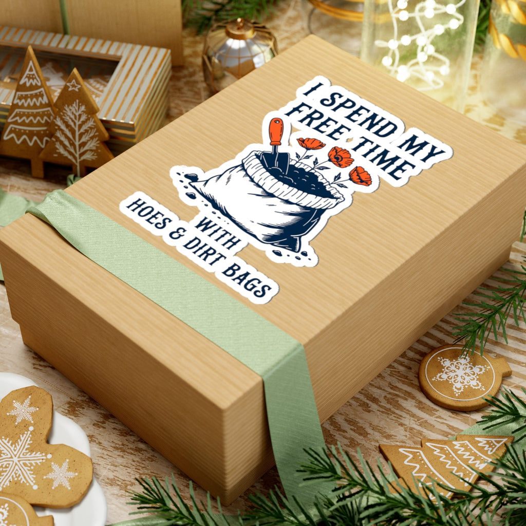 A light brown kraft gift box tied with a pale green ribbon, decorated with a humorous sticker that reads 'I SPEND MY FREE TIME WITH HOES & DIRT BAGS'. The box is styled on a rustic wooden table with Christmas cookies, pine branches, and holiday decorations.