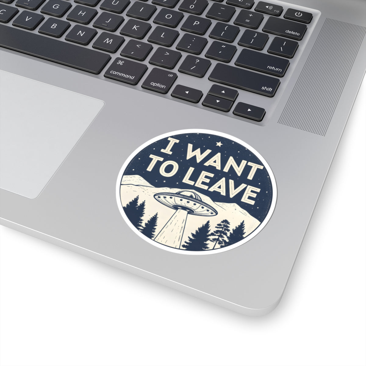 A round sticker with the text 'I WANT TO LEAVE' and an illustration of a UFO over a mountain landscape, shown applied to the lower part of a silver laptop keyboard.