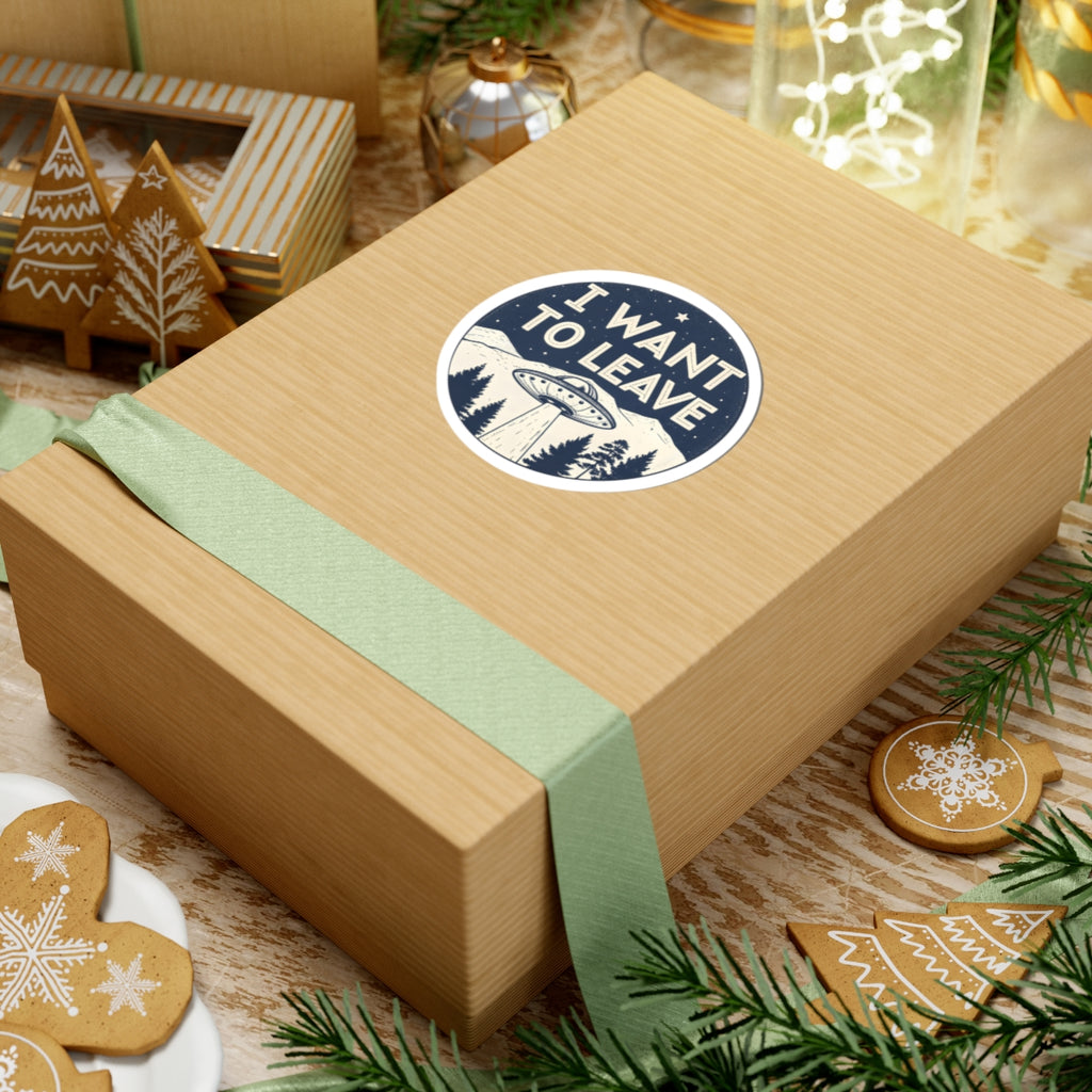 A round sticker with the text 'I WANT TO LEAVE' and an illustration of a UFO over a mountain landscape, applied to a wooden gift box. 