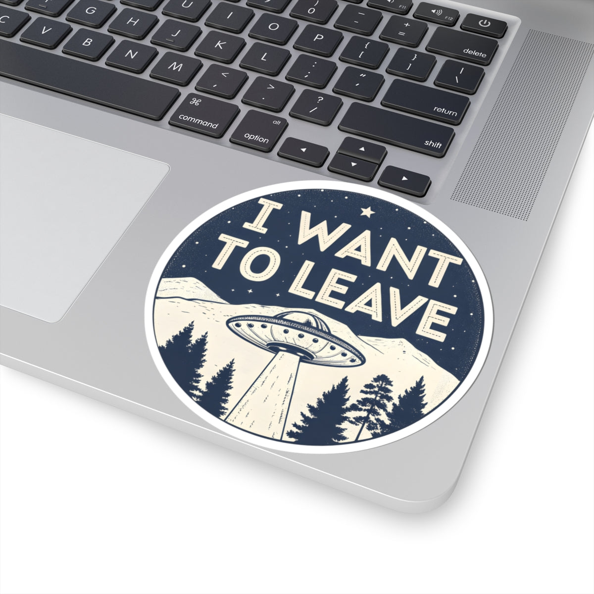 A round sticker with the text 'I WANT TO LEAVE' and a UFO flying over mountains and trees, shown applied to the corner of a silver laptop.