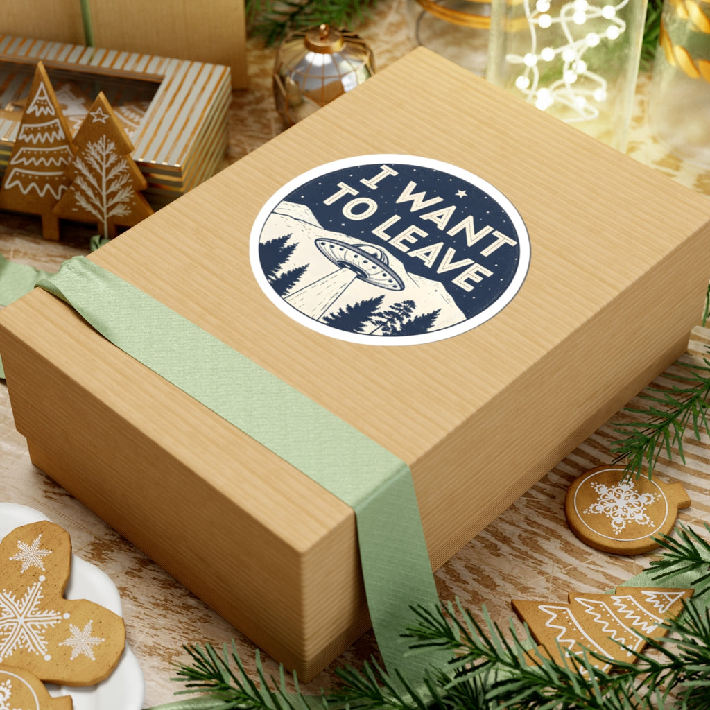 A square kraft paper gift box wrapped with a pale green ribbon, featuring a round sticker that reads 'I WANT TO LEAVE' with a UFO and mountain scene. The box is styled on a rustic table with holiday cookies, pine branches, and other gift boxes in the background.