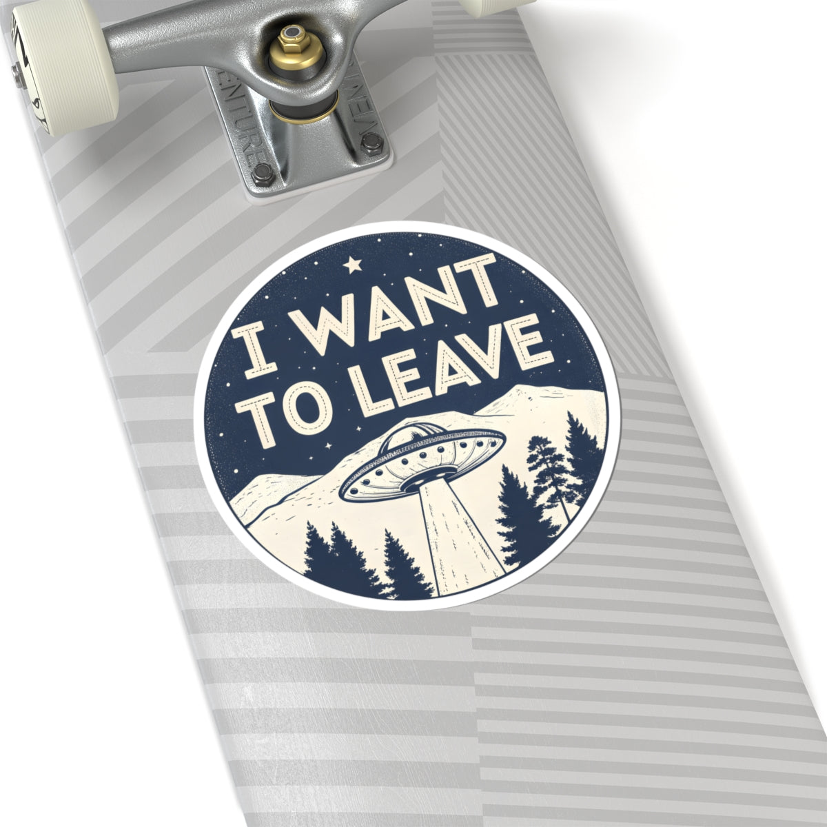 A circular sticker with the text 'I WANT TO LEAVE' and a UFO graphic, shown on the deck of a skateboard with gray striped grip tape.