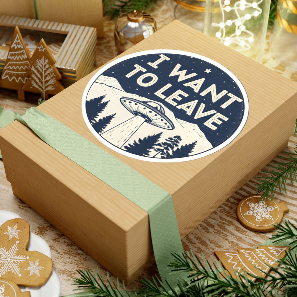 A square kraft gift box wrapped with a pale green ribbon, topped with a round sticker that reads “I WANT TO LEAVE” above a UFO and mountain scene. The box is styled on a tabletop with holiday cookies, evergreen branches, and other gift items in the background.