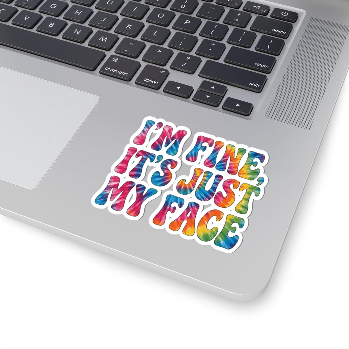 A colorful kiss-cut sticker with the text 'I'M FINE, IT'S JUST MY FACE' in a rainbow tie-dye pattern, shown applied to the lower right corner of a silver laptop.