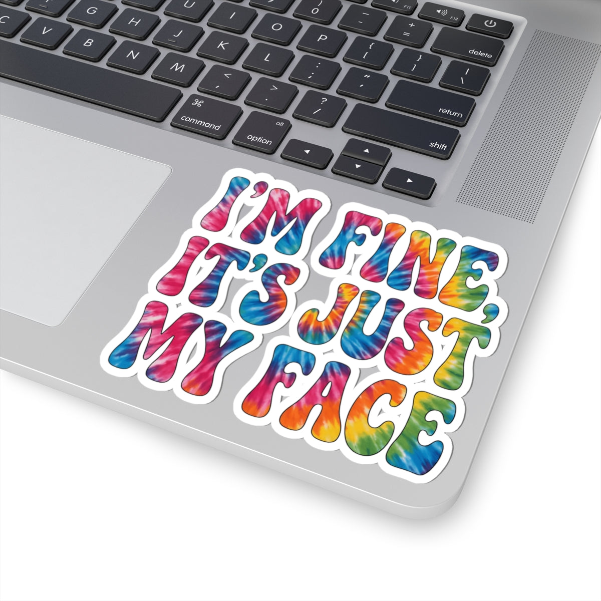 A kiss-cut vinyl sticker with the text 'I'M FINE, IT'S JUST MY FACE' in a colorful tie-dye pattern, shown applied to the corner of a silver laptop.