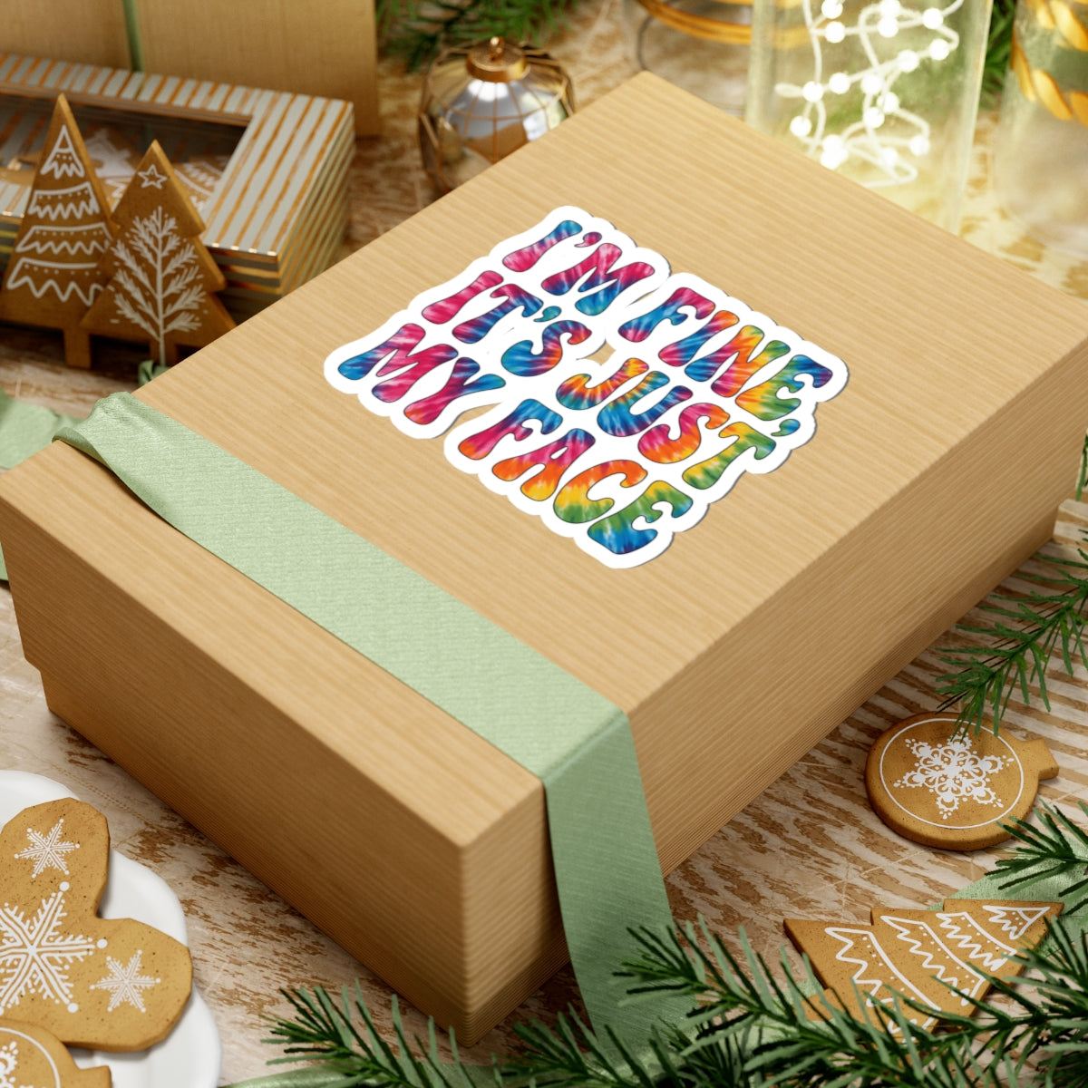 A kraft paper gift box wrapped with a pale green ribbon, featuring a colorful kiss-cut sticker that reads 'I'M SURE IT'S JUST MY FACE' in rainbow lettering. The box is styled on a rustic wooden surface with holiday cookies, evergreen branches, and other gift items in the background.