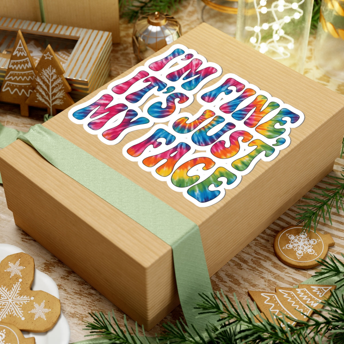 A square kraft paper gift box wrapped with a pale green ribbon, topped with a colorful kiss-cut sticker that reads “I’M FINE IT’S JUST MY FACE” in rainbow tie-dye lettering. The box is styled on a wooden surface with holiday decorations including gingerbread cookies, evergreen branches, and gift boxes in the background.
