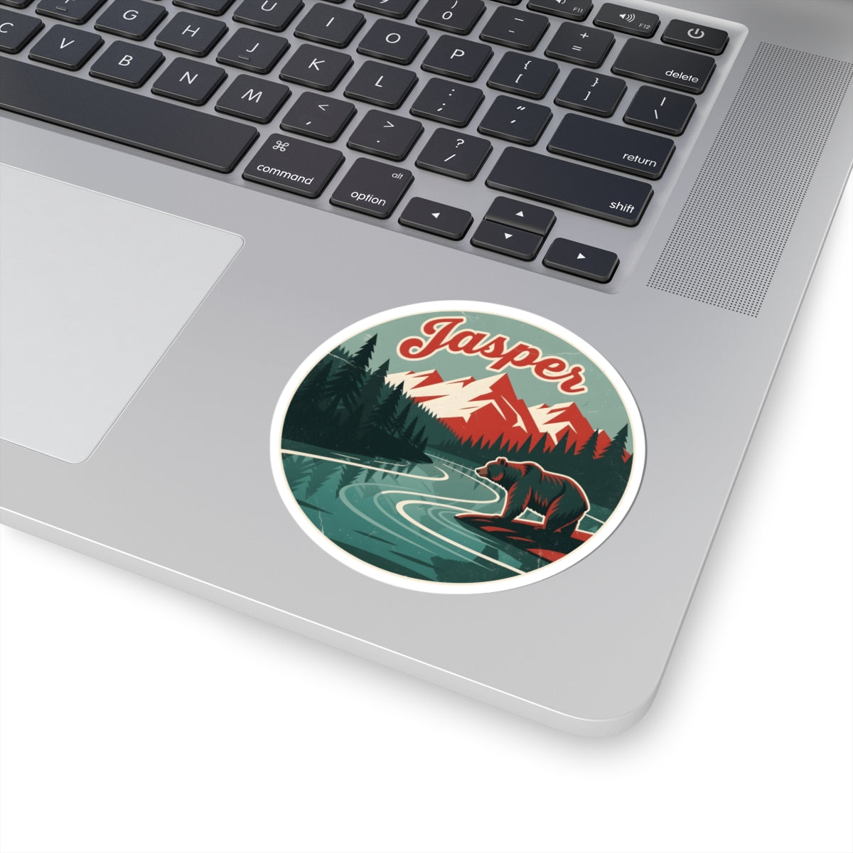 A round sticker with a retro-style illustration of a bear by a river, mountains, and the word 'Jasper' in red script, shown on the silver palm rest of a laptop keyboard.