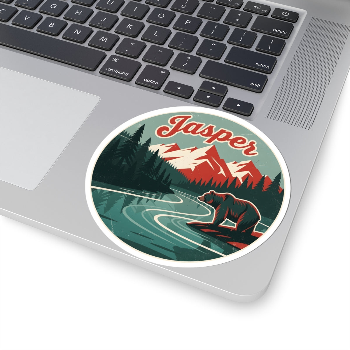 A round vinyl sticker with a retro-style illustration of a bear by a river and mountains, labeled 'Jasper', shown applied to the palm rest of a silver laptop.