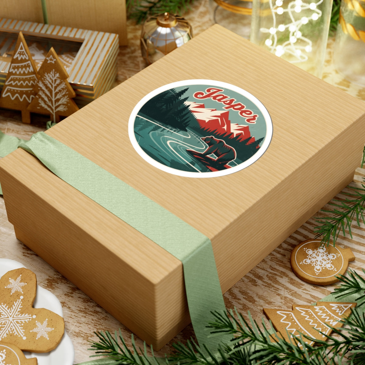 A square kraft paper gift box wrapped with a pale green ribbon, topped with a round sticker featuring a winter mountain scene and the word 'Jasper.' The box is styled on a tabletop with holiday cookies, evergreen branches, and other gift items in the background.