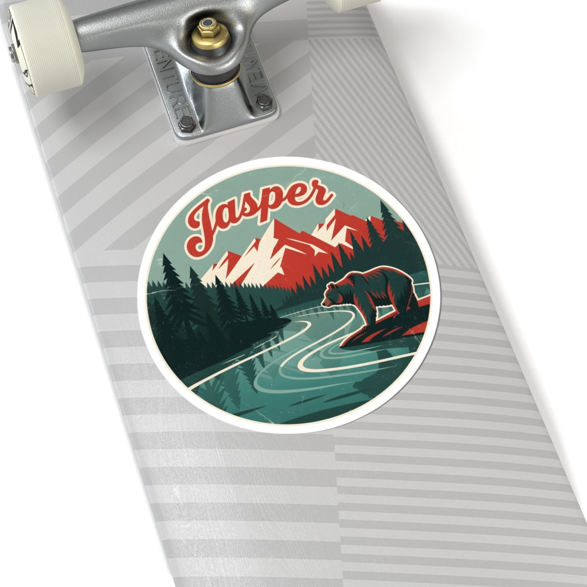 A close-up of a skateboard deck featuring a circular retro-style sticker with the word 'Jasper' and an illustration of a bear, mountains, trees, and a river. The skateboard has a gray and white striped pattern and is shown with part of the truck visible against a clean white background.