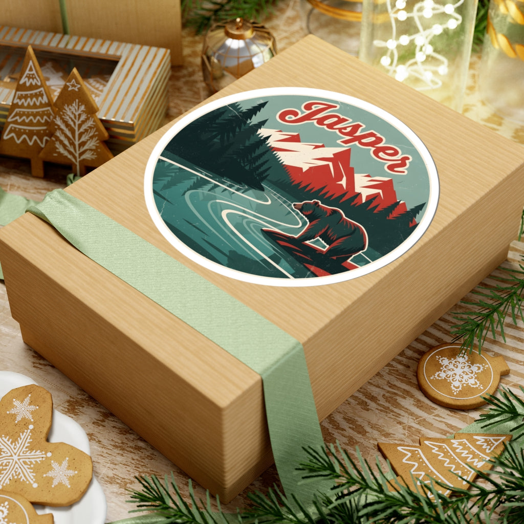 A square kraft gift box wrapped with a pale green ribbon and topped with a round vintage-style sticker reading "Jasper" over a mountain-and-river illustration, styled on a tabletop with holiday cookies, pine branches, and decor.