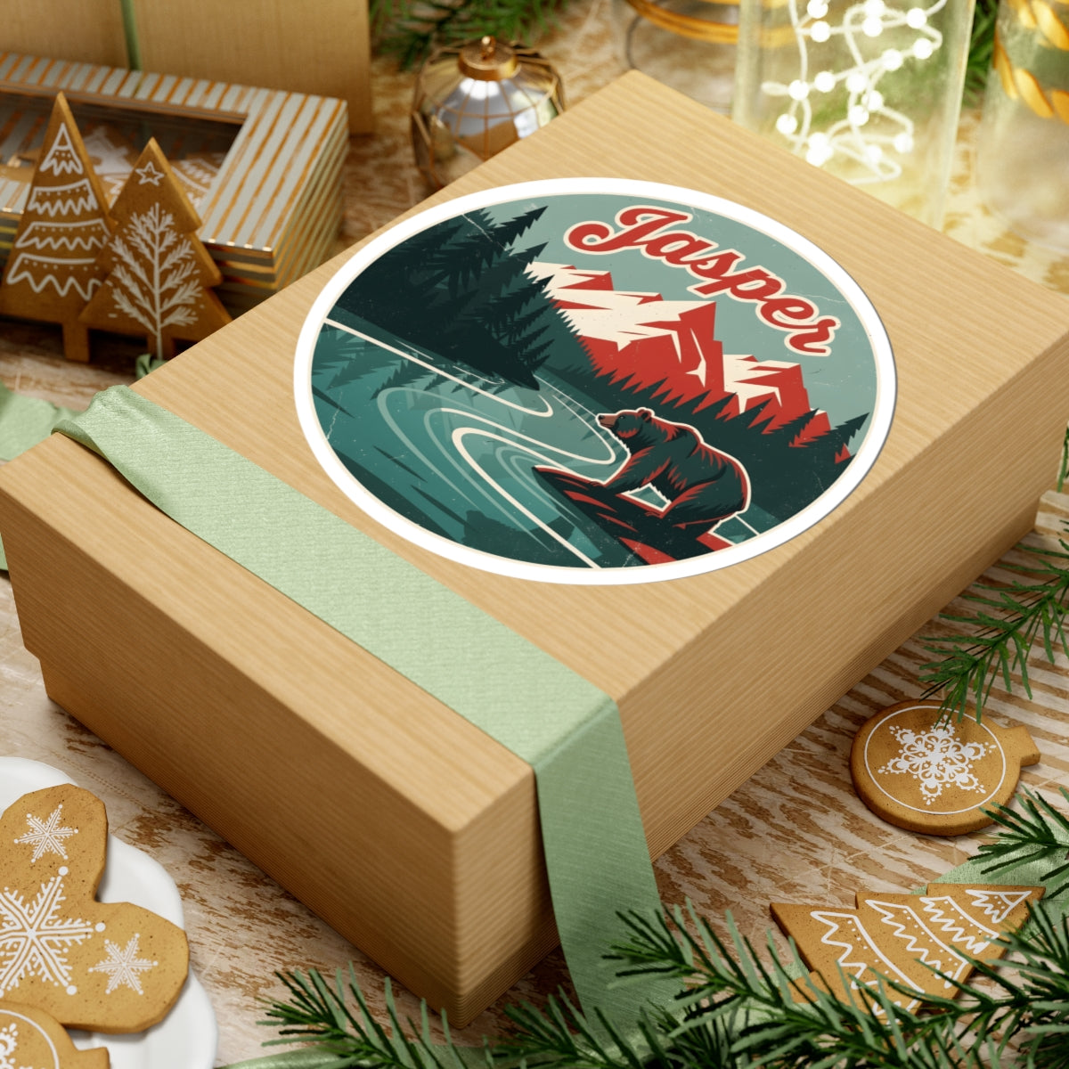 A square kraft gift box wrapped with a pale green ribbon and topped with a round vintage-style sticker reading "Jasper" over a mountain-and-river illustration, styled on a tabletop with holiday cookies, pine branches, and decor.