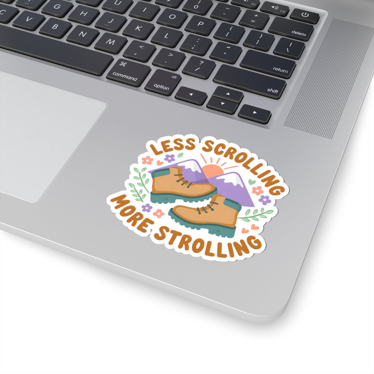 A kiss-cut vinyl sticker applied to the palm rest of a silver laptop. The sticker features an illustration of hiking boots, mountains, a sun, and flowers, surrounded by the text 'LESS SCROLLING MORE STROLLING' in orange lettering.