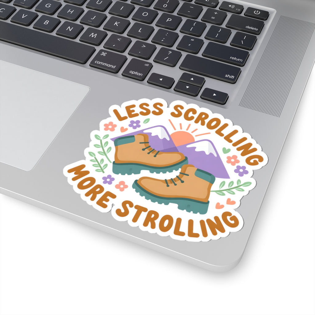 A kiss-cut vinyl sticker with the text 'LESS SCROLLING MORE STROLLING' and an illustration of hiking boots, mountains, flowers, and a sun, shown applied to the corner of a silver laptop.