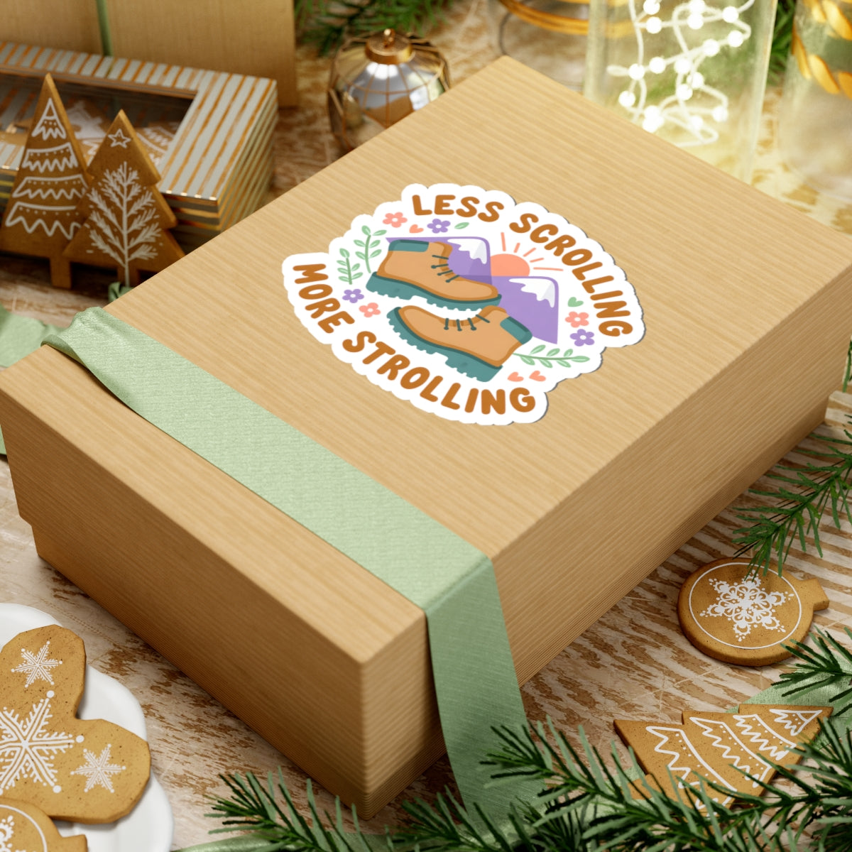 A square kraft paper gift box tied with a mint green ribbon, featuring a kiss-cut sticker that reads 'LESS SCROLLING MORE STROLLING' with an illustration of a pair of boots and scenic background. The box is styled on a wooden surface with Christmas cookies, pine branches, and holiday decorations.