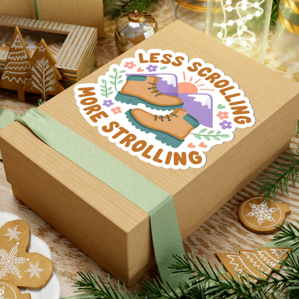 A square kraft gift box wrapped with a pale green ribbon, featuring a kiss-cut sticker that reads 'LESS SCROLLING MORE STROLLING' with an illustration of tan hiking boots, purple mountains, and small flowers. The box is styled on a wooden surface with holiday cookies, evergreen branches, and warm decorative lights in the background.