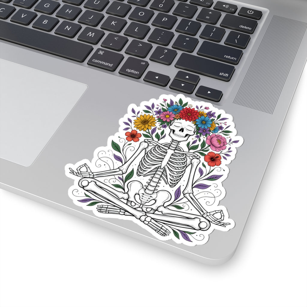 A kiss-cut vinyl sticker of a black and white skeleton meditating amidst colorful flowers, shown applied to the silver surface of a laptop next to the keyboard.