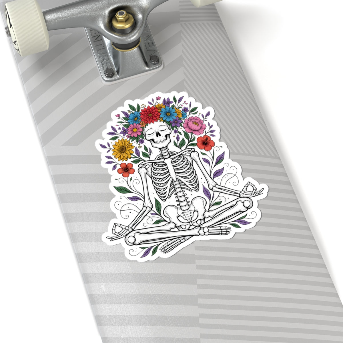 A kiss-cut sticker featuring a Day of the Dead skeleton sitting cross-legged under a colorful flower crown, shown applied to the deck of a skateboard.