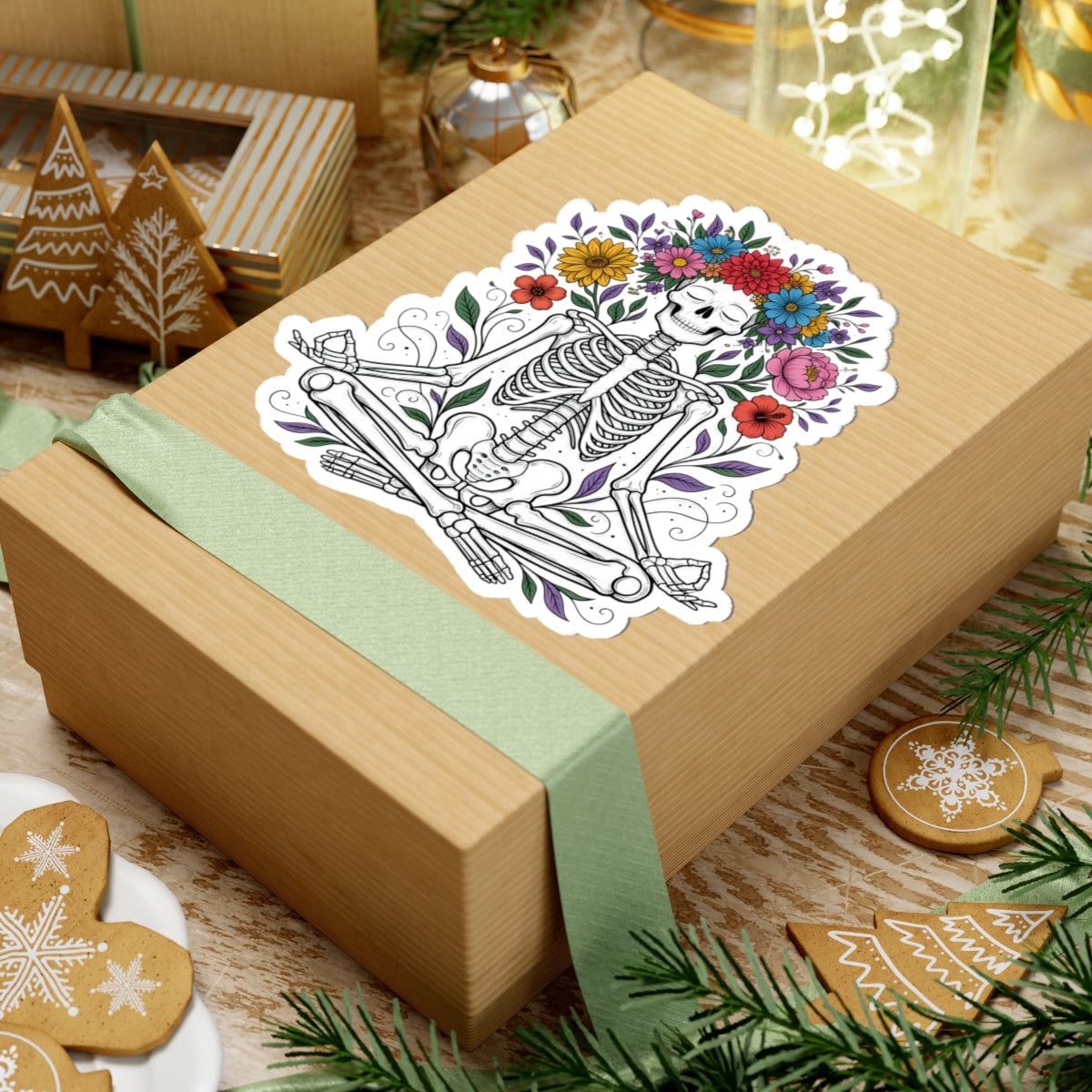 A kraft paper gift box wrapped with a pale green ribbon, topped with a kiss-cut sticker featuring a skeleton surrounded by colorful flowers. The box is styled on a rustic wooden surface with holiday cookies, evergreen branches, and other gift items in the background.