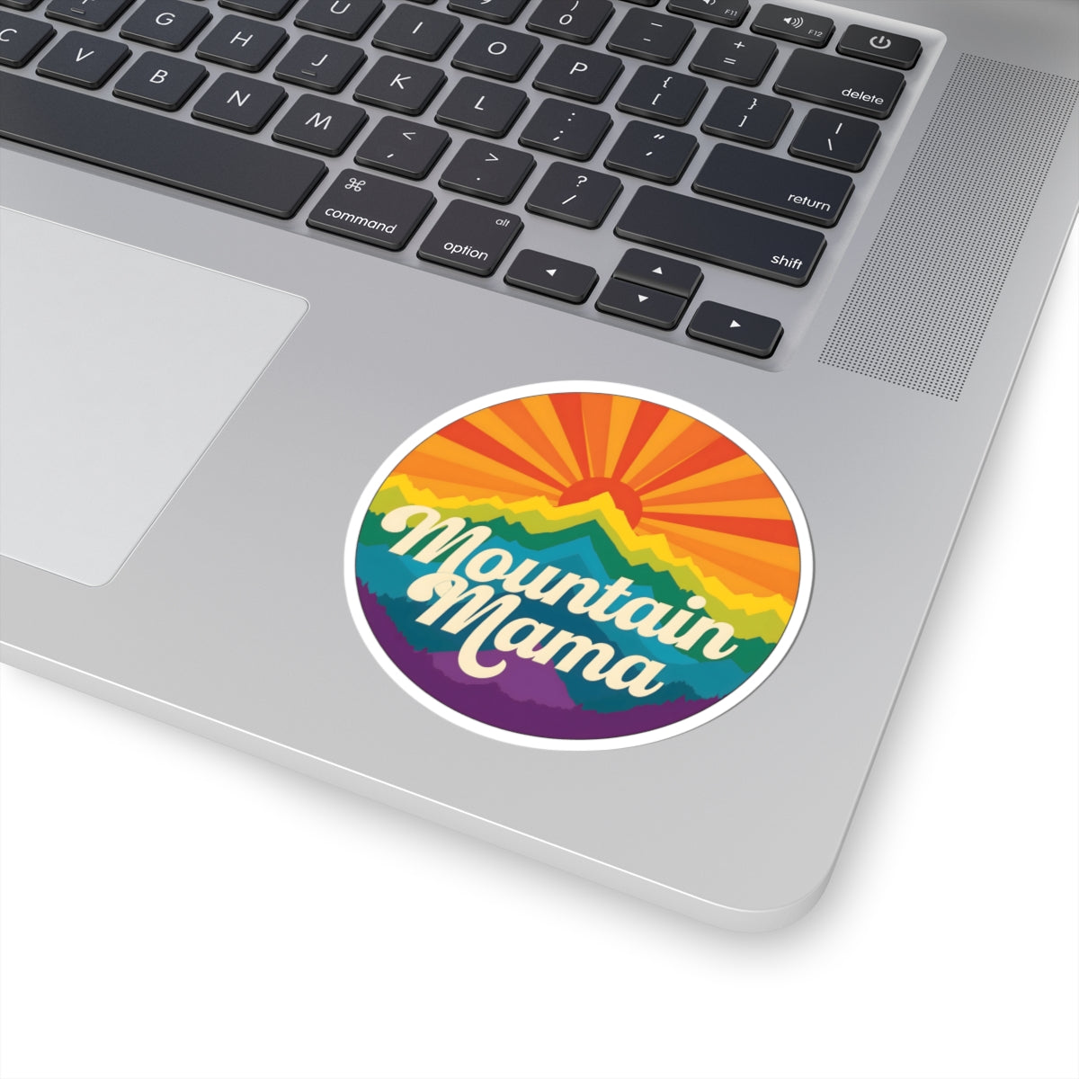 A round vinyl sticker with the text 'Mountain Mama' in white script over a colorful rainbow mountain sunset design, shown applied to the silver palm rest of a laptop keyboard.
