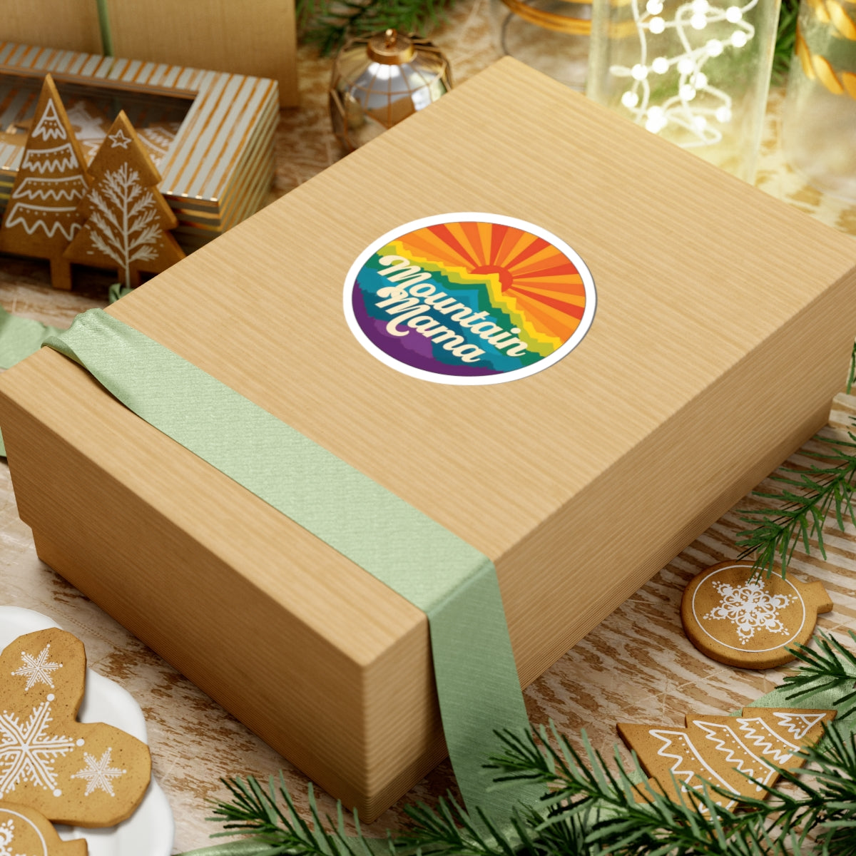 Wooden gift box with a round 'Mountain Mama' mountain and sunset sticker on a festive background with cookies and lights.