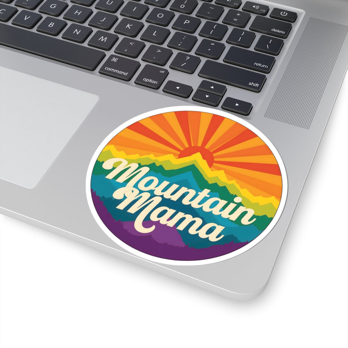 A round vinyl sticker with a retro mountain sunset design and the text 'Mountain Mama' in white script, shown applied to the corner of a silver laptop.