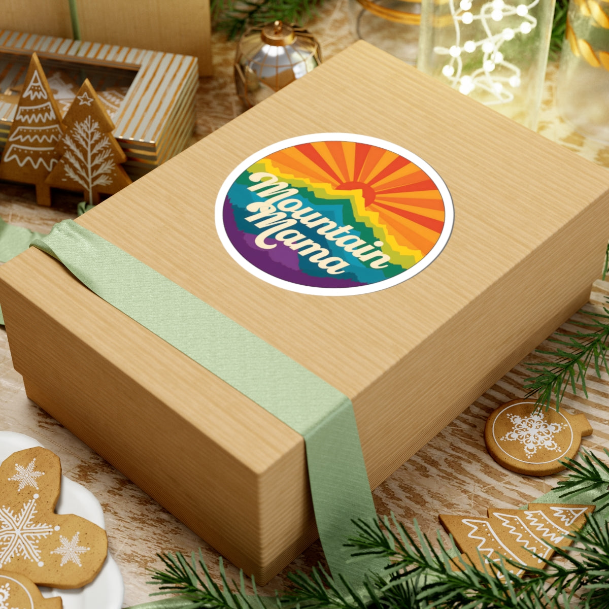 A kraft paper gift box wrapped with a pale green ribbon and topped with a round sticker that reads “Mountain Mama” over a rainbow sunburst, styled on a tabletop with holiday cookies, pine branches, and glass decor.