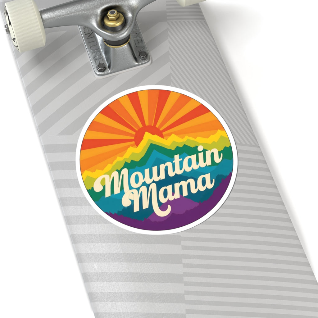 A circular sticker reading 'Mountain Mama' in white script over a retro rainbow mountain design, shown on a gray striped skateboard deck.