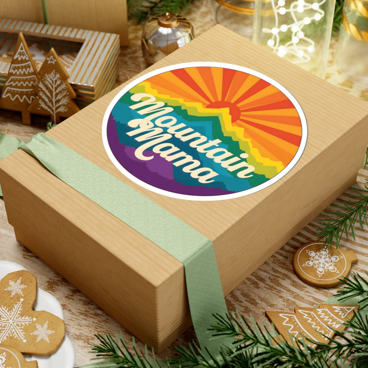A kraft paper gift box wrapped with a pale green ribbon, topped with a round sticker that reads “Mountain Mama” in white script over a rainbow sunburst design. The box is styled on a rustic table with holiday cookies, pine branches, and other gift items.