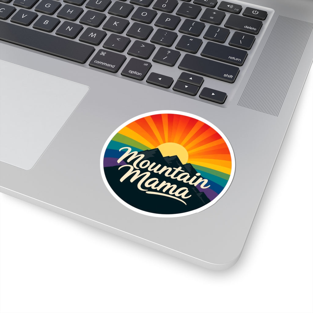 A round sticker with the text 'Mountain Mama' and a colorful sunset mountain design, shown applied to the palm rest of a silver laptop.