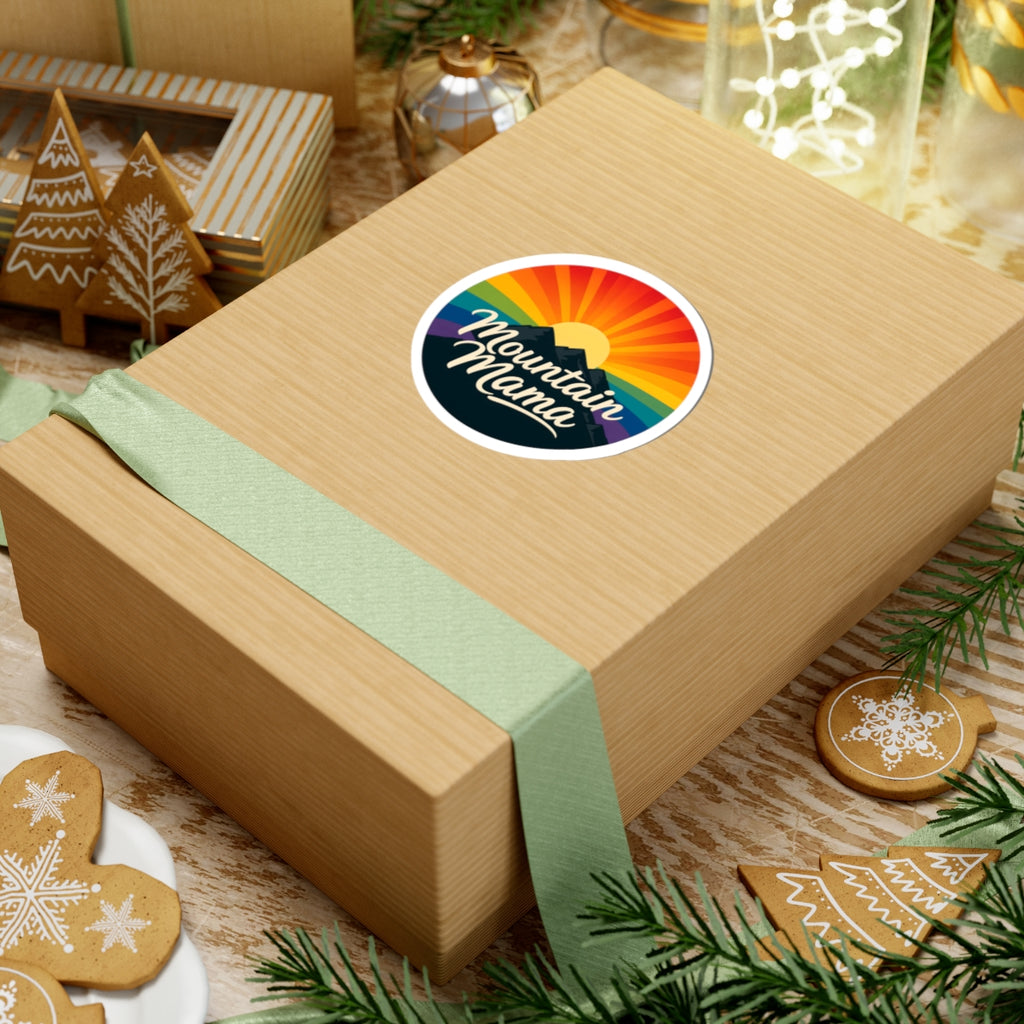 A square kraft paper gift box wrapped with a pale green ribbon and topped with a round sticker that reads "Mountain Maine" over a rainbow sunset, styled on a tabletop with holiday cookies, pine branches, and other gift items.