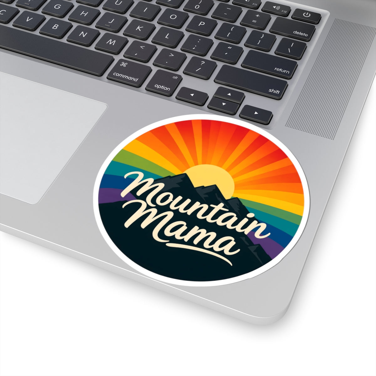 A round vinyl sticker with a retro mountain sunset design and the words 'Mountain Mama' in white script, shown applied to the palm rest of a silver laptop.