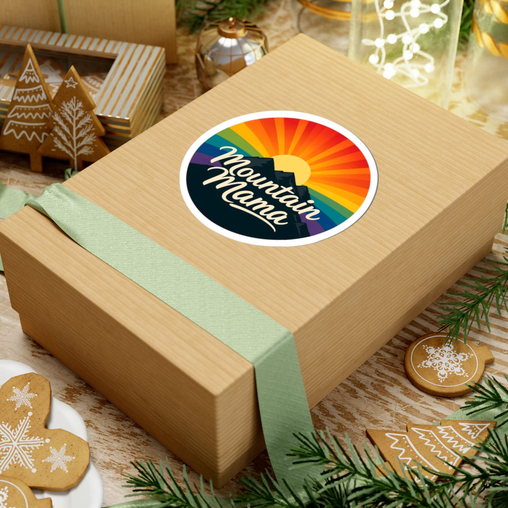 A kraft paper gift box tied with a pale green ribbon, featuring a round sticker that reads 'Mountain Mama' over a rainbow sunset and mountain graphic. The box is styled on a rustic table with holiday cookies, pine branches, and other gift items in the background.