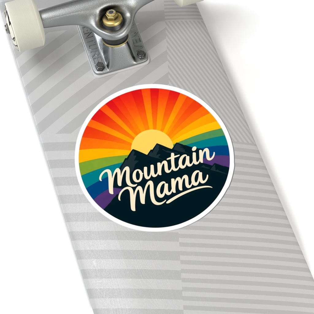 A circular sticker with a retro rainbow sunburst, mountain silhouette, and the words 'Mountain Mama' in white script, shown applied to the deck of a skateboard.
