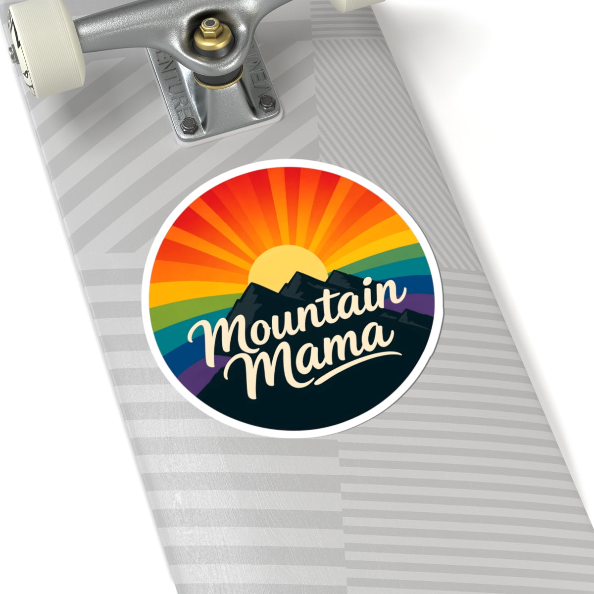 A circular sticker with a retro rainbow sunburst, mountain silhouette, and the words 'Mountain Mama' in white script, shown applied to the deck of a skateboard.