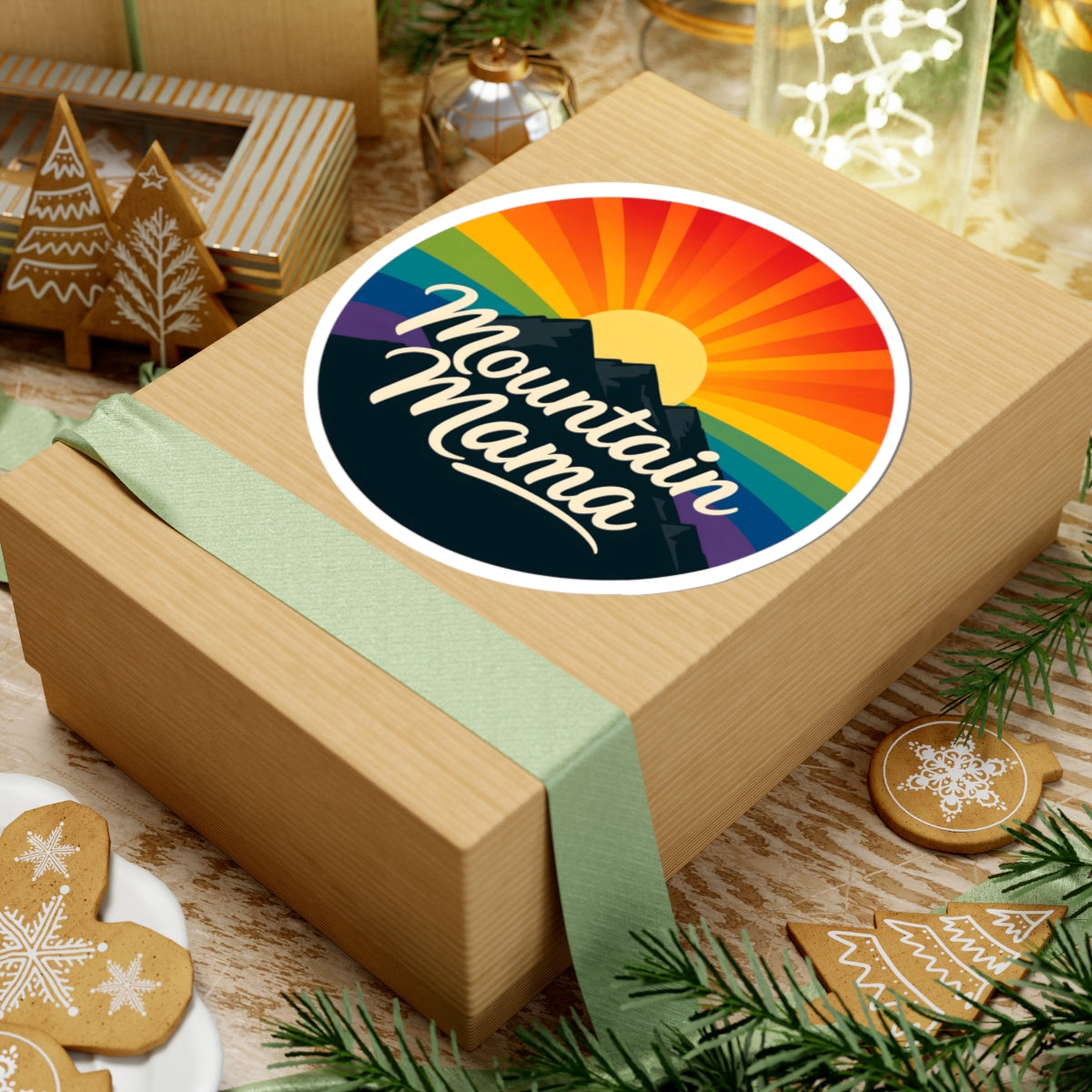 A round sticker with a colorful mountain sunset design and the words 'Mountain Mama' is shown on top of a kraft gift box wrapped with a pale green ribbon. The scene is styled with holiday cookies, pine branches, and other gift items.