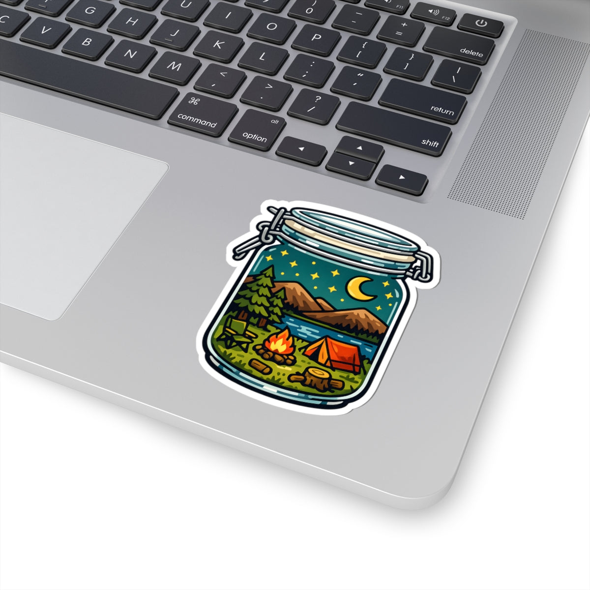 A kiss-cut vinyl sticker of a camping scene inside a glass jar, shown applied to the corner of a silver laptop keyboard.
