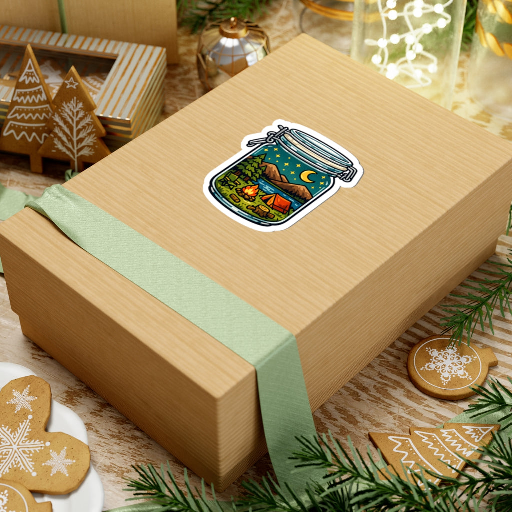 A kraft paper gift box wrapped with a pale green ribbon, featuring a kiss-cut sticker of a mason jar filled with camping scenery on top. The box is styled on a rustic wooden surface with Christmas cookies, pine branches, and other gift boxes in the background.