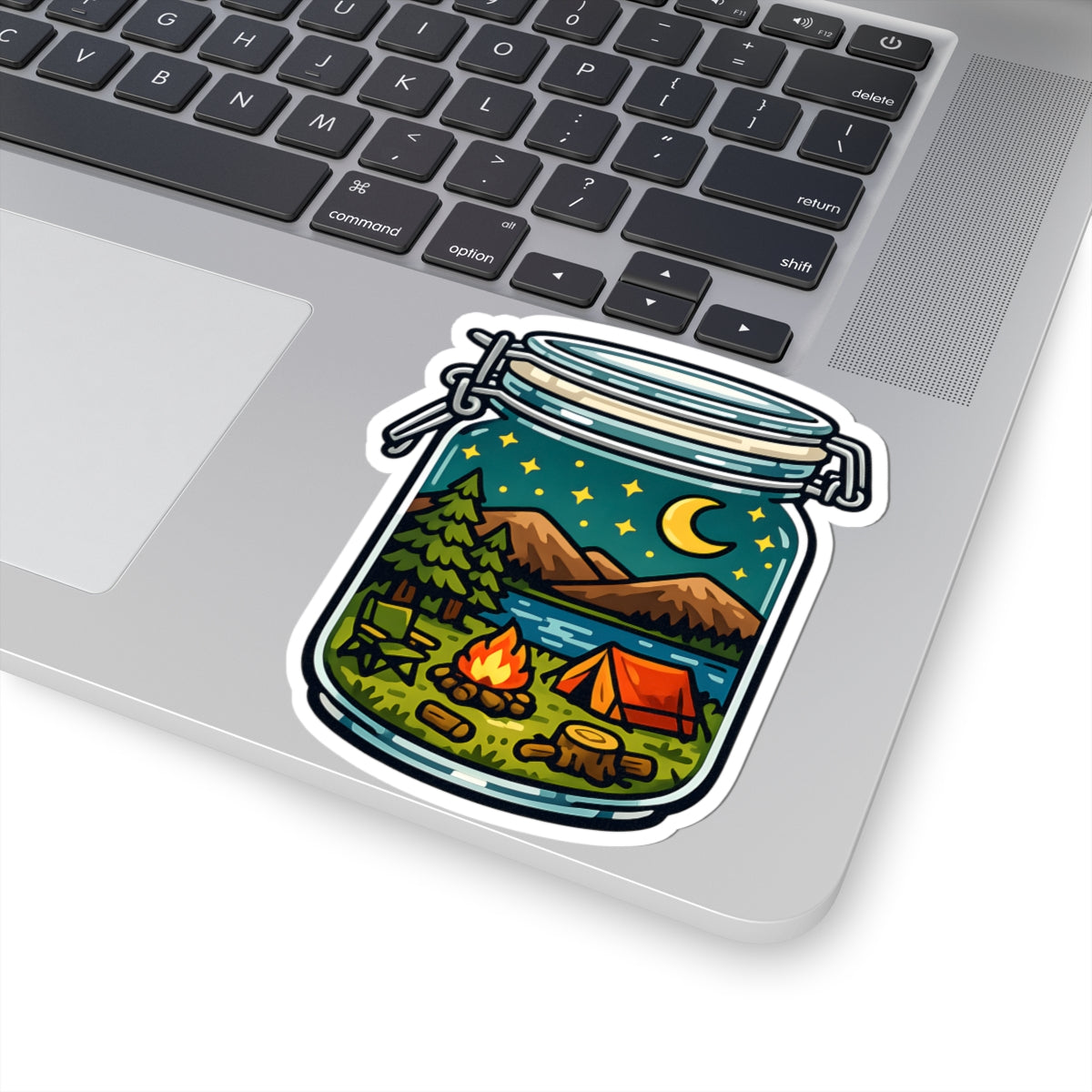 A kiss-cut vinyl sticker of a camping scene inside a mason jar, shown applied to the corner of a silver laptop. The sticker features a detailed illustration of a campsite with a tent, campfire, trees, mountains, and a starry night sky.