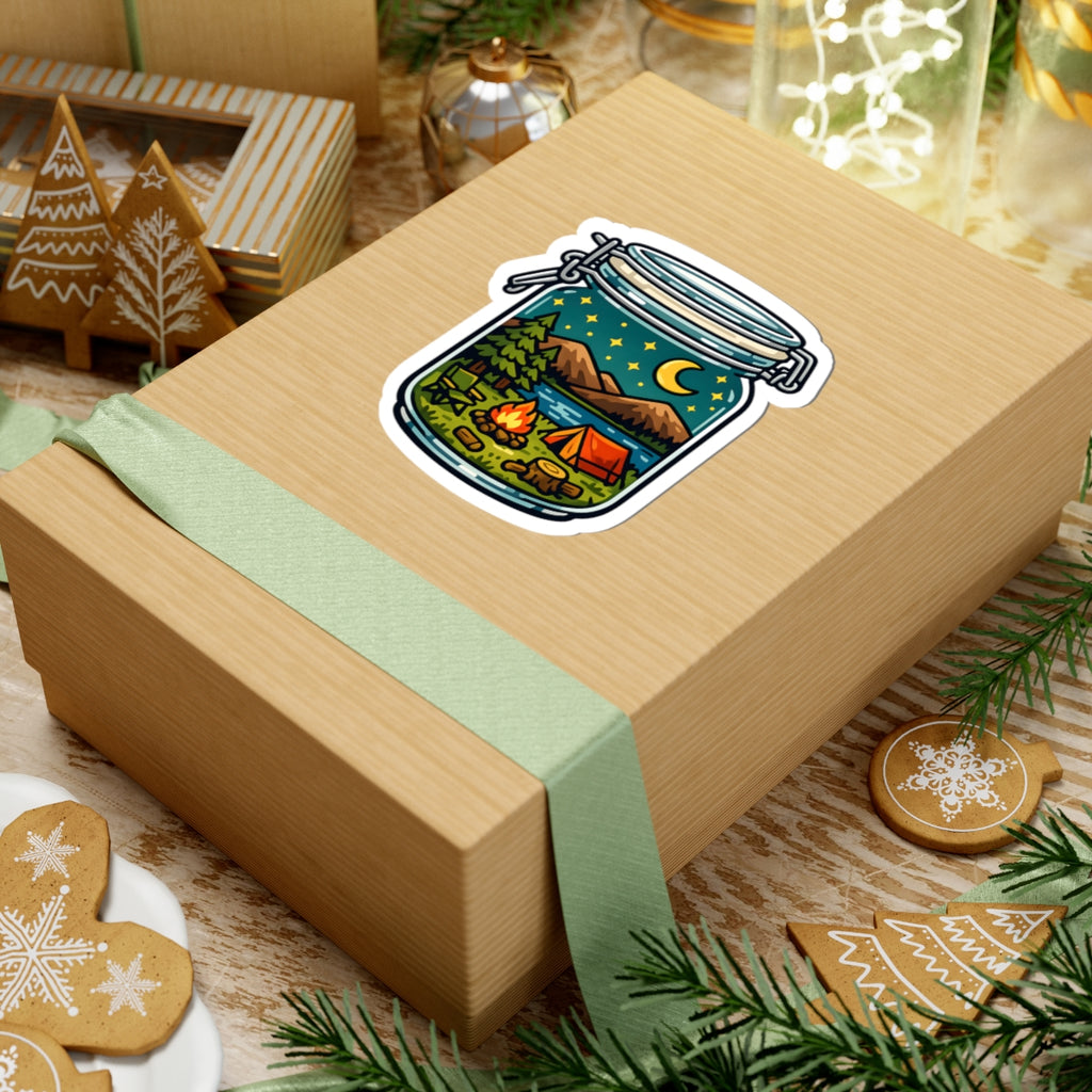 A square kraft gift box wrapped with a pale green ribbon, featuring a kiss-cut sticker of a camping scene inside a mason jar on top. The box is styled on a wooden surface with holiday cookies, pine branches, and other gift items in the background.