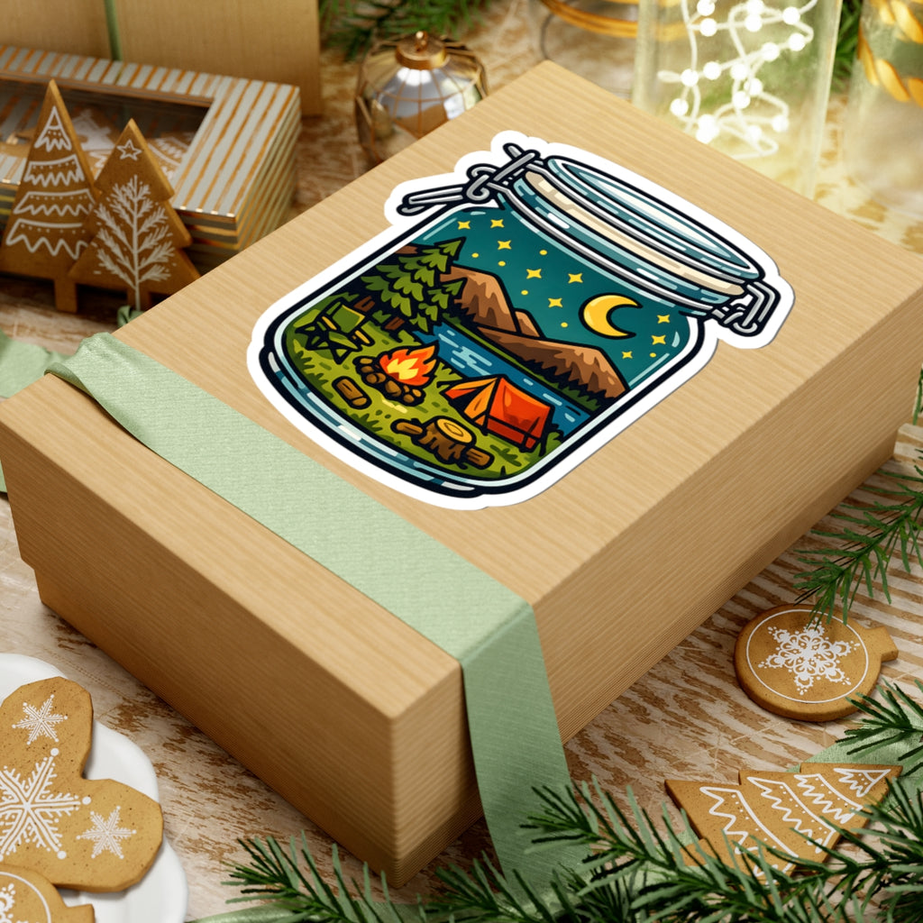 A square kraft gift box wrapped with a pale green ribbon, featuring a kiss-cut sticker of a camping scene inside a mason jar on top. The box is styled with holiday cookies, pine branches, and warm festive background decor.
