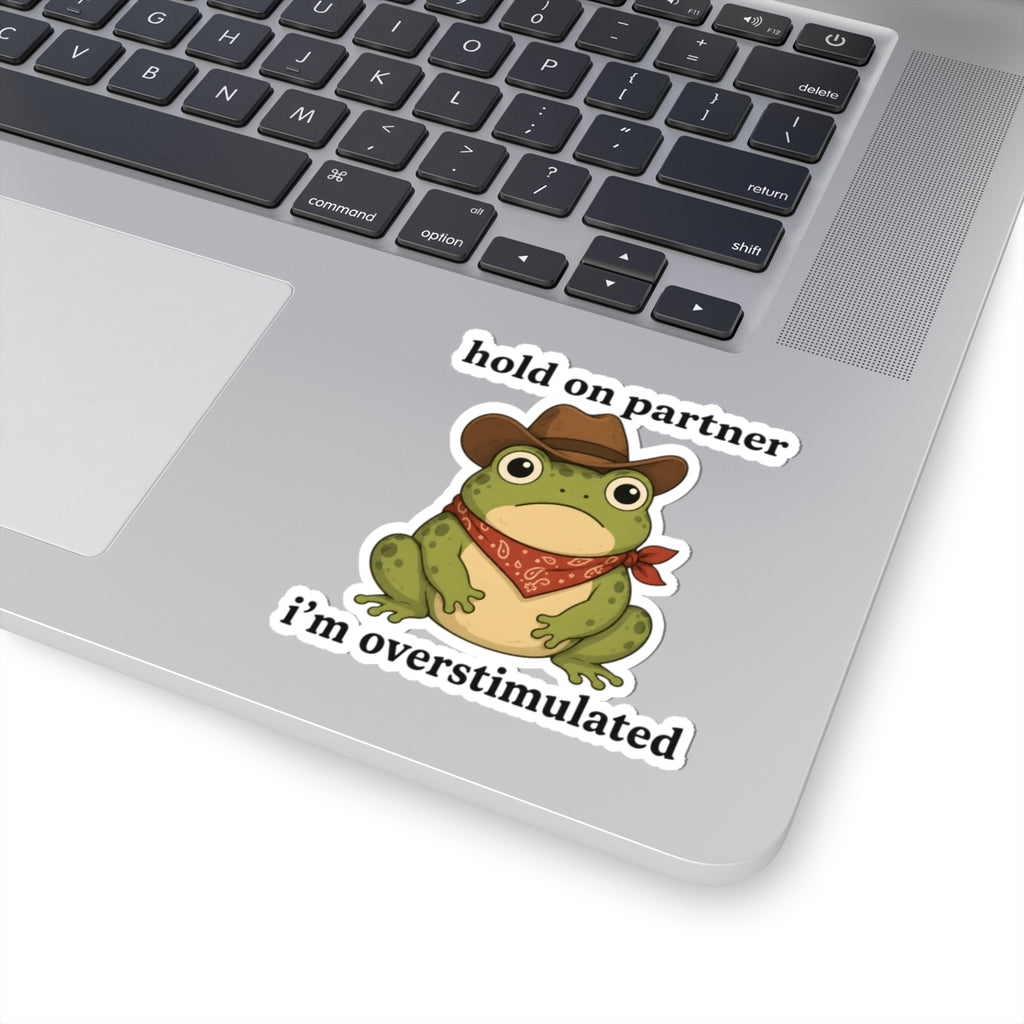 A kiss-cut vinyl sticker featuring a cartoon green frog wearing a brown cowboy hat and red bandana, with the text 'hold on partner i'm overstimulated', shown applied to the lower right corner of a silver laptop.