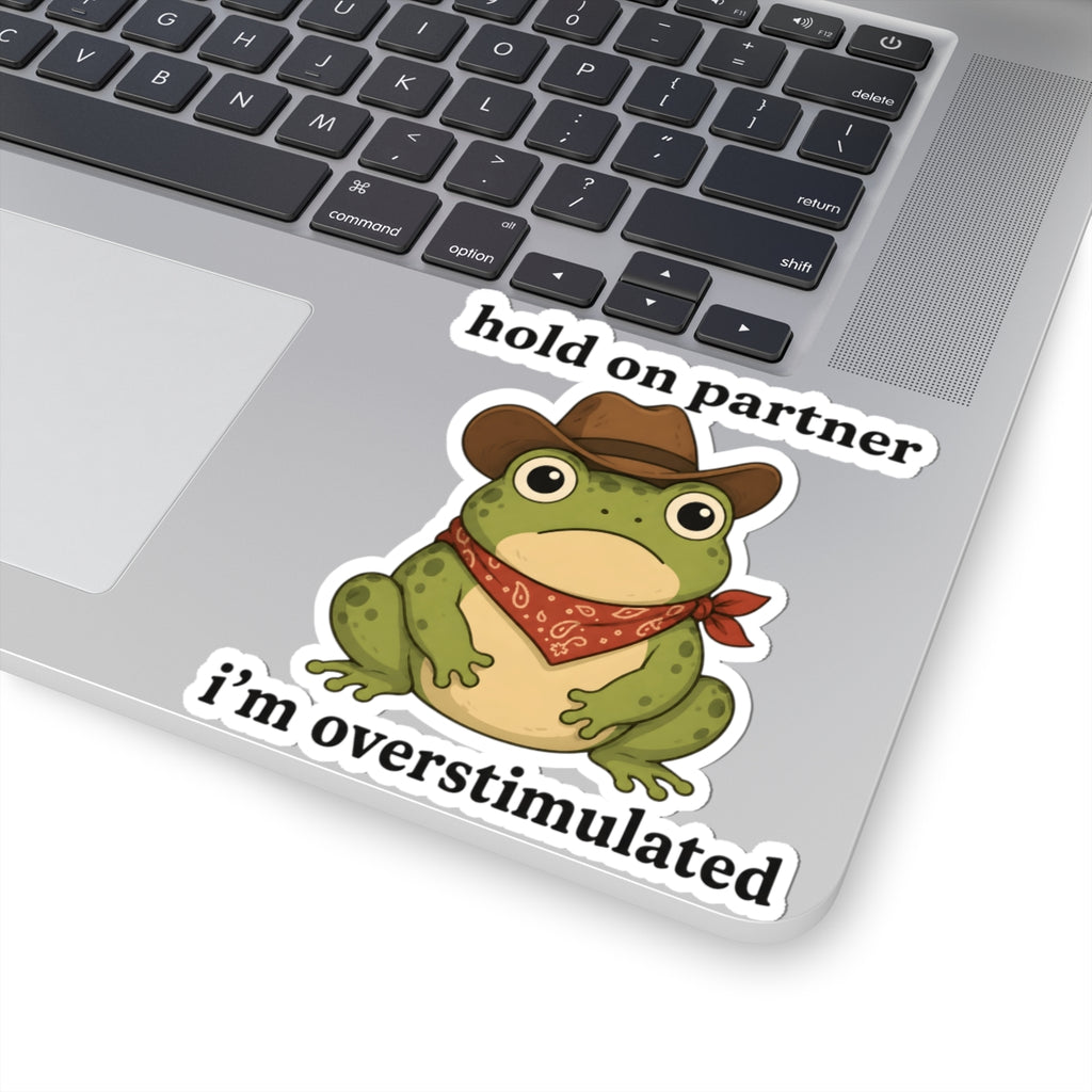 A kiss-cut vinyl sticker featuring a cartoon green frog wearing a brown cowboy hat and red bandana, with the text 'hold on partner i'm overstimulated'. The sticker is shown applied to the silver surface of a laptop keyboard.
