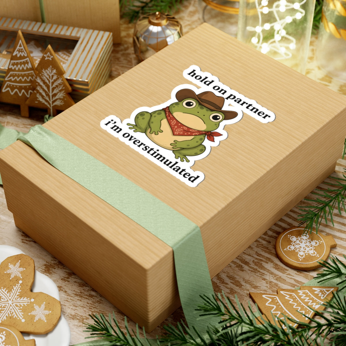 A square kraft paper gift box tied with a pale green ribbon, featuring a kiss-cut sticker of a green cartoon frog wearing a cowboy hat and bandana with the text 'hold on partner I'm overstimulated'. The box is styled on a rustic wooden surface with holiday decorations including gingerbread cookies, pine branches, and a small lantern.