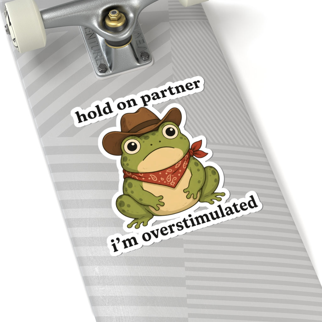 A kiss-cut sticker featuring a cartoon green frog wearing a brown cowboy hat and red bandana, with the text 'hold on partner i'm overstimulated' in white outlined lettering. The sticker is shown applied to the gray striped surface of a skateboard deck, with part of the silver truck and white wheels visible.