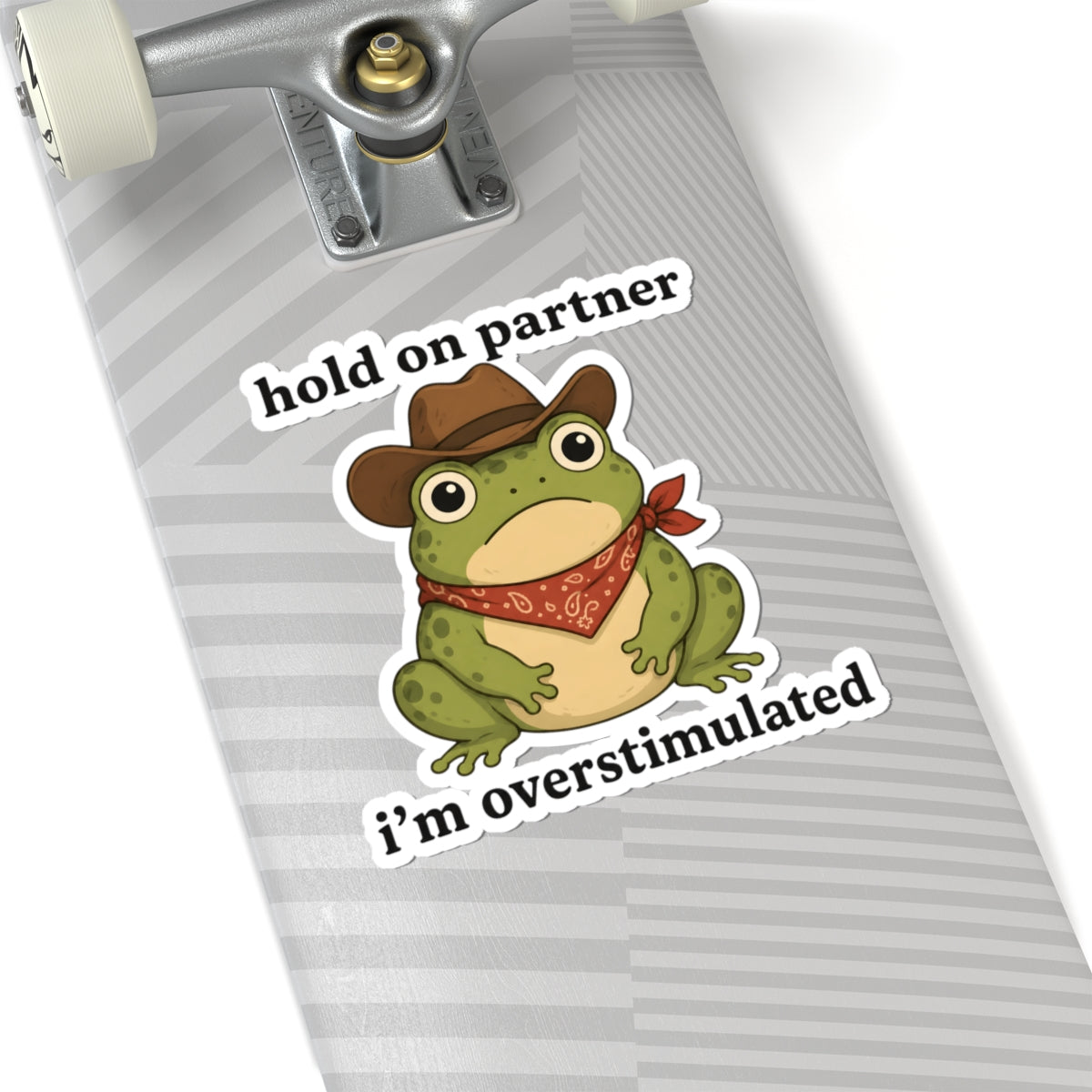 A kiss-cut sticker featuring a cartoon green frog wearing a brown cowboy hat and red bandana, with the text 'hold on partner i'm overstimulated' in white outlined lettering. The sticker is shown applied to the gray striped surface of a skateboard deck, with part of the silver truck and white wheels visible.